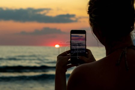 Photo for Silhouette of a woman at the beach photographing the sunset with a smatphone. Large copy space and twilight tones. Unrecognizable back side - Royalty Free Image