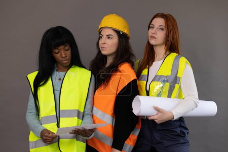 Foto de Tres modelos de diferentes etnias posando, vestidos como trabajadores de la construcción. Chalecos reflectantes, sombreros duros y proyecto enrollado. - Imagen libre de derechos