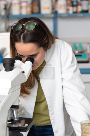 Foto de Mujer científica joven bajo un microscopio en un laboratorio científico. Concepto de investigación para enfermedades raras y excelencia científica. Biólogo durante una prueba de laboratorio con reactivos en el fondo - Imagen libre de derechos