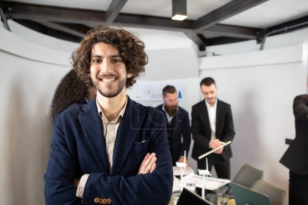Téléchargez les photos : Jeune homme d'affaires beau sourire dans un bureau. Portrait posé avec un accent sélectif sur l'homme d'affaires et les collègues flous en arrière-plan - en image libre de droit