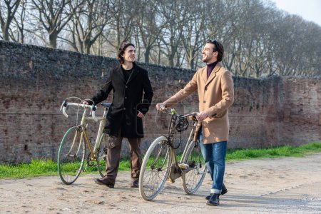 Two stylish men stand with their vintage bicycles, engaging in a lively conversation on a sunlit path with old brick walls and bare trees in the background