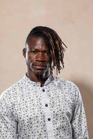 Relaxed young man with dreadlocks standing against a neutral background in a printed shirt, reflecting modern style and casual confidence