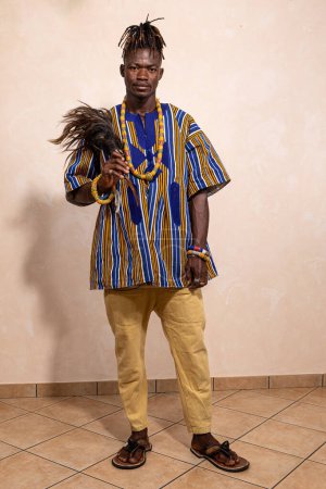Fier homme africain présentant des vêtements traditionnels vibrants avec une plume cérémonielle, célébrant le riche patrimoine culturel et l'identité