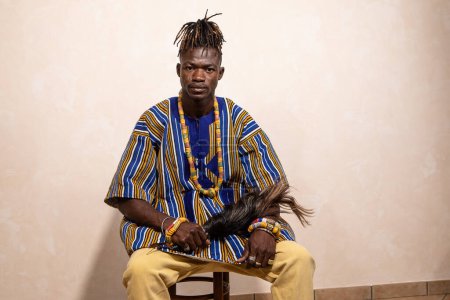 Ein sitzender afrikanischer Mann in farbenfroher traditioneller Tracht, der eine Feder in der Hand hält, repräsentiert kulturelles Erbe und zeitlose Tradition in einer modernen Umgebung