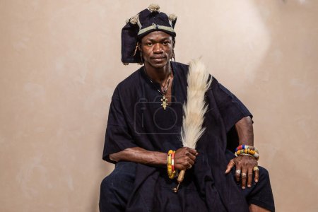 Portrait d'un homme africain vêtu d'un habit cérémonial traditionnel, avec ornements et accessoires culturels. Il tient fièrement un bâton à plumes tribales, symbolisant le patrimoine culturel et l'identité