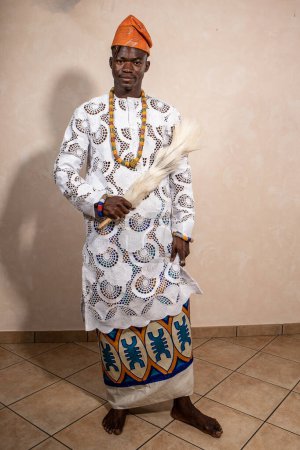 An African man exudes elegance in a traditional white outfit with intricate patterns, complemented by a vibrant tribal hat. His attire reflects a blend of modern style and rich cultural heritage