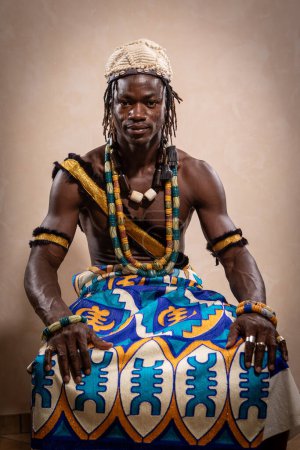 A skilled African drummer poses confidently in vibrant traditional clothing, adorned with symbolic jewelry. He holds a cultural drum, ready to perform rhythmic beats that tell stories of his ancestry