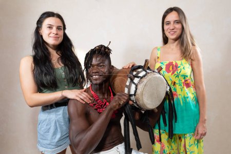 Zwei lächelnde Frauen und ein Mann mit traditioneller Trommel zelebrieren kulturelle Vielfalt mit einem lebhaften Gruppenporträt, das Freude und Einheit zeigt