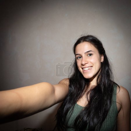 Eine fröhliche junge Frau mit langen dunklen Haaren fängt einen glücklichen Moment mit einem Selfie im Haus ein und zeigt ihre natürliche Schönheit und ihr ansteckendes Lächeln