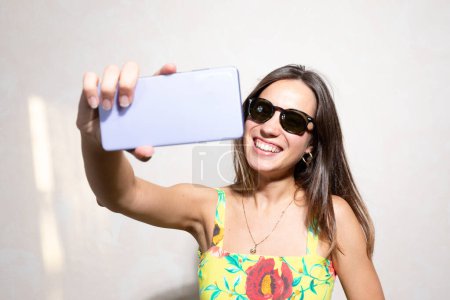 A cheerful young woman with long hair and sunglasses captures a joyful moment with her smartphone, taking a selfie while wearing a colorful summer dress against a neutral backdrop