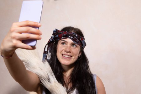 A vivacious young woman adorned with a stylish headscarf smiles as she captures a selfie using her smartphone, exuding a sense of joy and contemporary fashion