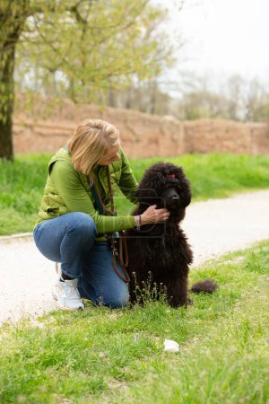 In a moment of affection, a woman kneels down on a park path to hug her loyal black poodle, surrounded by the greenery of early spring