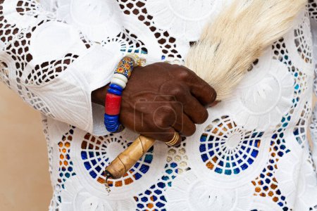 Close-up view of an African person's hand wearing traditional clothing, showcasing elaborate embroidery and holding an animal tail, complemented by vibrant beaded bracelets