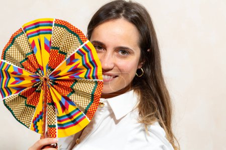 A vibrant portrait of a smiling woman playfully covering half of her face with a brightly colored African print fan. Her white shirt provides a striking contrast to the fan's vivid pattern, while her simple jewelry complements the festive atmosphere