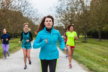 An active group of women in various colors of sportswear, from blue to neon green, run together along a park's gravel pathway, enjoying the camaraderie and challenge of group exercise