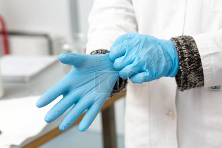A medical professional is shown wearing a white coat and meticulously putting on sterile blue gloves in a clinical laboratory setting, highlighting the importance of hygiene
