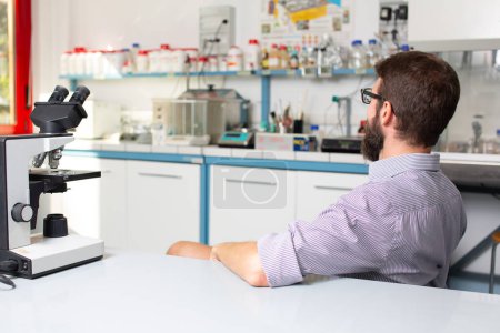 A male scientist in a striped shirt sits thoughtfully in a lab environment, contemplating his research with a microscope awaiting
