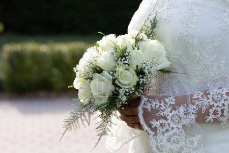 eine zarte Nahaufnahme fängt den spitzenärmeligen Arm einer Braut ein, der einen Strauß weißer Rosen umschlingt, was Eleganz und eine zeitlose Hochzeitstradition symbolisiert
