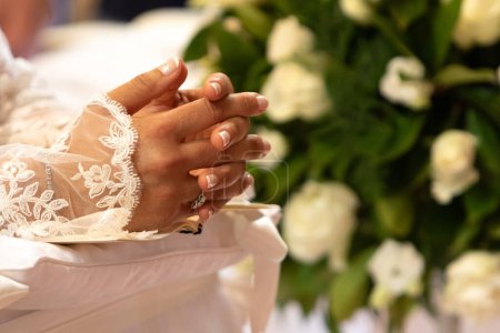 Die Hände einer Braut, geschmückt mit einem zarten Ehering, sind im Gebet über ihr spitzenverziertes Kleid gefaltet, im Hintergrund weiße Rosen