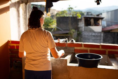 une femme est immergée dans la routine du lavage des vêtements à la main, debout dans un bassin en béton sur un toit baigné de lumière du soleil