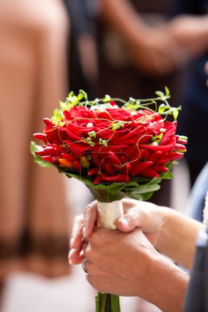ein einzigartiger Hochzeitsstrauß aus roten Rosen und lebendigen Chilischoten, den jemand in der Feier fest in der Hand hält