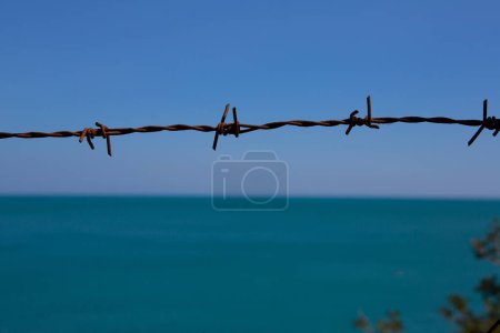 Téléchargez les photos : Clôture en fil de fer barbelé rouillé s'étend à travers, avec un fond bleu océan serein suggérant contraste et frontières - en image libre de droit