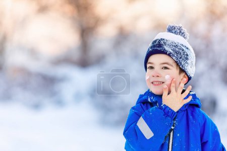 Foto de Retrato de niño sonriente aplicando crema fría a la cara al aire libre. Concepto de protección de piel invierno - Imagen libre de derechos