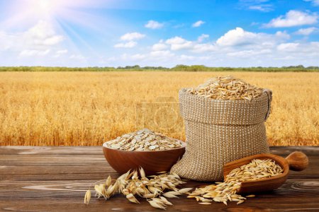 Photo for Uncooked oatmeal in bowl and oat grains in burlap bag on table with ripe cereal field on the background - Royalty Free Image