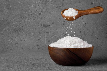 sea salt crystals falling from wooden spoon in bowl on grey background