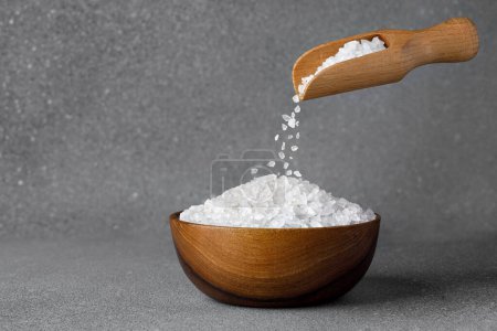 Photo for Sea salt crystals falling from wooden scoop in bowl on grey background - Royalty Free Image