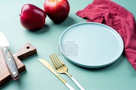 Foto de Clean empty plate mockup, green plate with cutlery and red apples and napkin on green table, table setting. Space for text or menu.Business food brand template. - Imagen libre de derechos