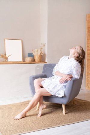 Foto de Hermosa mujer caucásica en vestido de moda de verano blanco sentado en una silla suave descalzo, mirando hacia otro lado. Boho o scandi interior - Imagen libre de derechos