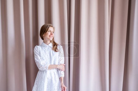 Photo for Beautiful caucasian woman in white summer fashion dress standing on beige curtain background, looking away. Pastel colors - Royalty Free Image