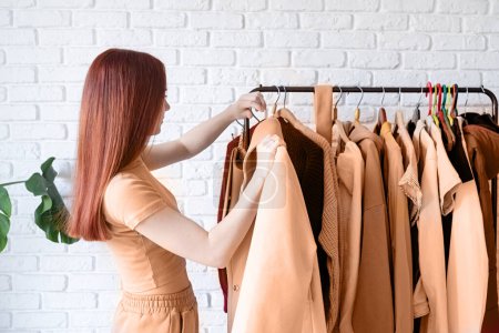 Farbe des Jahres 2024: Peach Fuzz. schöne junge Frau in beigem Mantel, die vor Kleiderbügel steht und Outfit wählt. Auswahl eines Kleiderschranks, Stylisten, Einkaufen. Bekleidung entwerfen