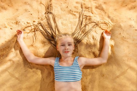  A smiling girl with braids lies on the sand in a striped swimsuit, happy and bathed in sunlight.