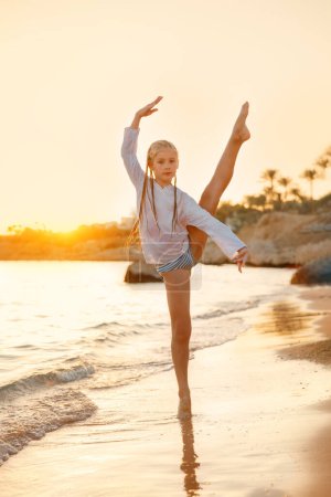 Ein Mädchen tanzt bei Sonnenuntergang am Strand. Sie steht barfuß im Sand, ein Bein hoch, die Hände anmutig erhoben..