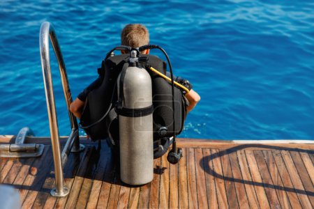 A teenage boy in a diving suit with an oxygen tank on his back getting ready to dive underwater on the wooden deck of a yacht.