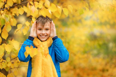 A teenager in a blue jacket and yellow scarf playing hide and seek behind a yellow leaf against the backdrop of autumn nature.