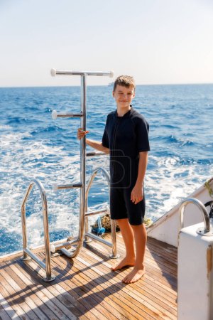 A 14-year-old boy in a diving suit stands on the stern of a yacht, looking at the camera and smiling. The sea is visible in the background, and the sunlight makes the picture bright and cheerful. 