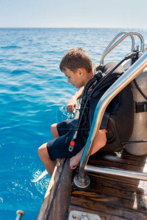 A teenager sits on the stern of the yacht, dressed in a diving suit. He has an oxygen tank over his shoulder. He is looking at the water and preparing to dive.