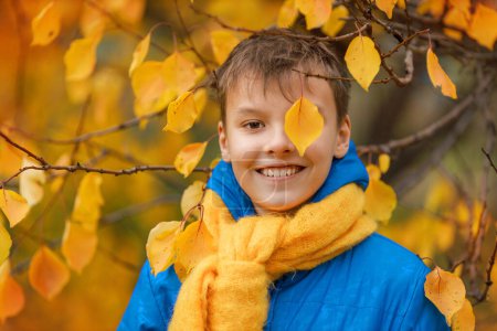 Ein Teenager in blauer Jacke und gelbem Schal spielt Verstecken hinter einem gelben Blatt vor dem Hintergrund der herbstlichen Natur.