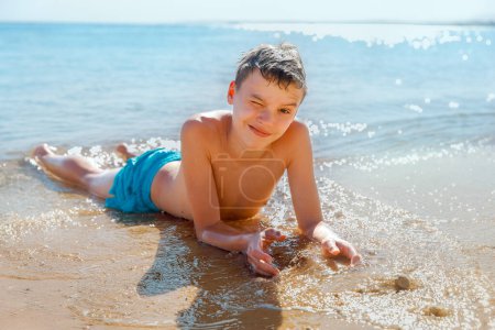 A teenager lies on the sand by the sea. He is wearing blue swimming shorts. The bright sun shines, and behind it, there is a view of the sea.