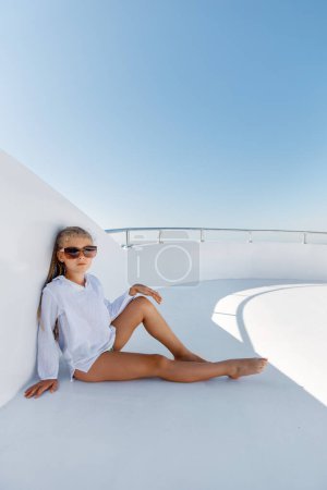 A girl in sunglasses and a white tunic is sitting on the roof of a yacht sailing on the sea.