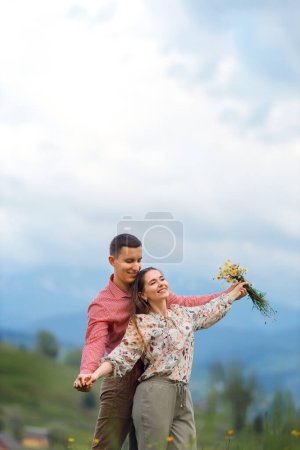 A romantic couple standing against a backdrop of beautiful sky and mountains. The man stands behind with both of them extending their arms as if theyre flying. The woman holds a bouquet of flowers.