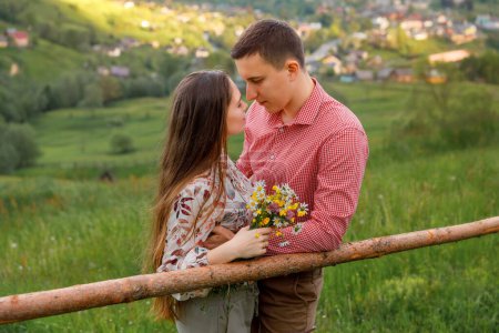 A young couple is standing face to face, leaning against a wooden beam. Behind them is nature and spring greenery. The woman is holding a bouquet, and they are gazing at each other tenderly.