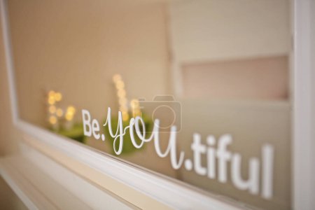 "Be You Tiful" cutting sticker words on the mirror. Blurred reflection of bedroom interior. Combination symbol of Be Your Self and You Are Beautiful motivational quotes for women. Self love concept.