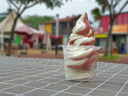 Soft Serve Ice Cream White Vanilla mix Chocolate swirl in a clear plastic cup on an outdoor table. Ice Cream sundae on daylight, close up view, selective focus with blurred outdoor background.