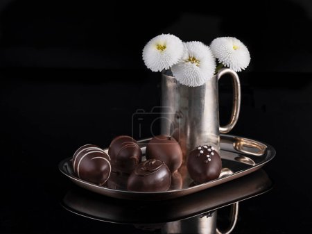 Foto de Chocolates preciosos bellamente dispuestos en bandeja de plata, florero vintage decorado de plata con un ramo de flores de margarita. - Imagen libre de derechos