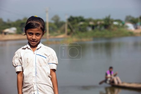 Photo for Taherpur, Bangladesh - November 05, 2019: Little girl standing in front of the river wearing a school dress. Poor school-going girl wearing a torn dress is ready to go to school. - Royalty Free Image