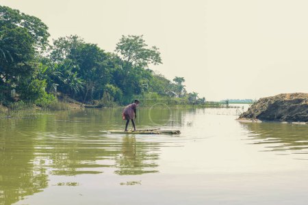 Téléchargez les photos : Taherpur, Bangladesh - 05 novembre 2019 : Un petit garçon dérive d'un radeau en pilotant une pagaie pendant l'inondation dans un village isolé. Petit garçon ramant radeau artisanal ou bateau fait de bananiers attachés ensemble. - en image libre de droit